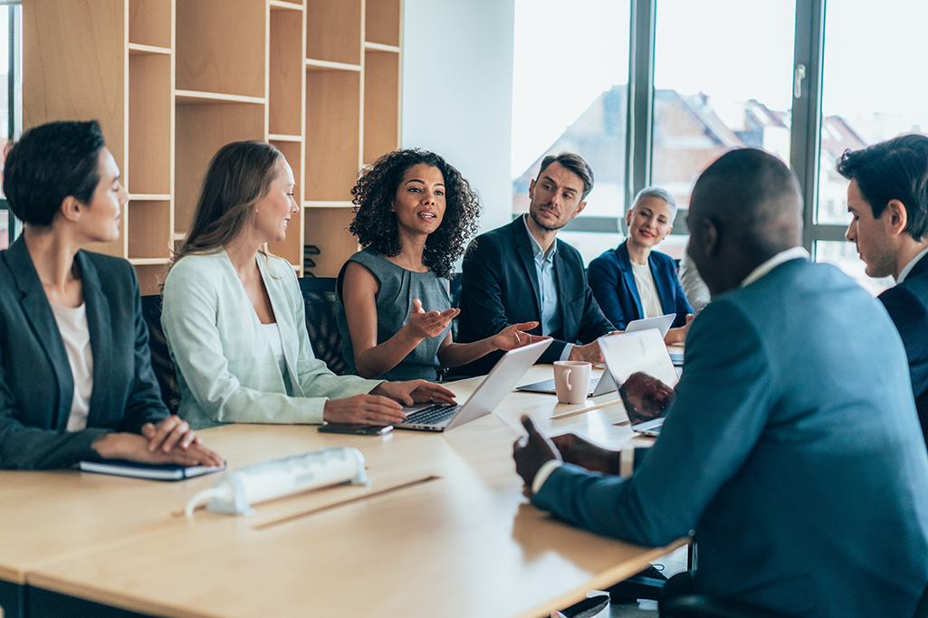 Group of seven professionals of diverse background sitting at a board table. They are engaged in discussion