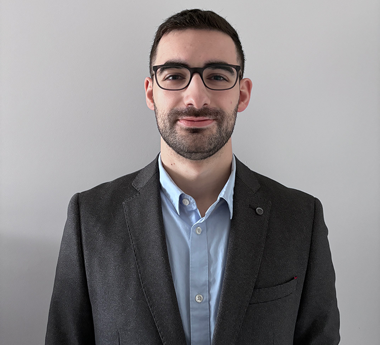 Louai Bibi headshot. He is wearing a dark grey suit jacket and a light blue dress shirt. He has a short beard and dark glasses