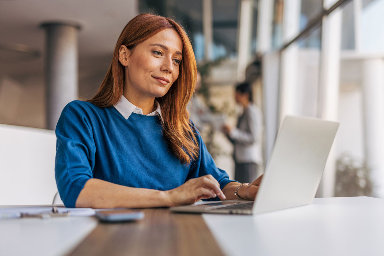Woman with red hair and blue sweater looking over a laptop in an open office