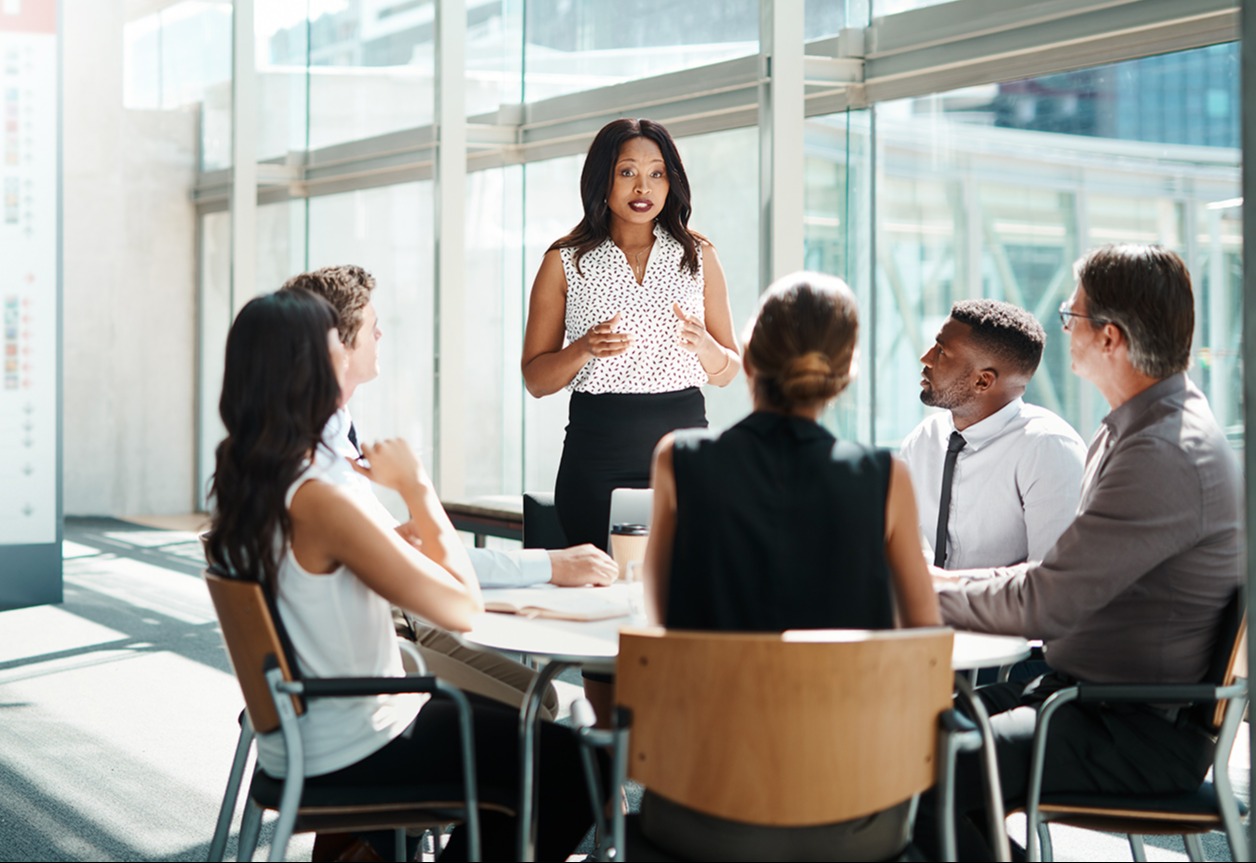 A woman standing in front of a group of people at a table in an office setting explains a concept